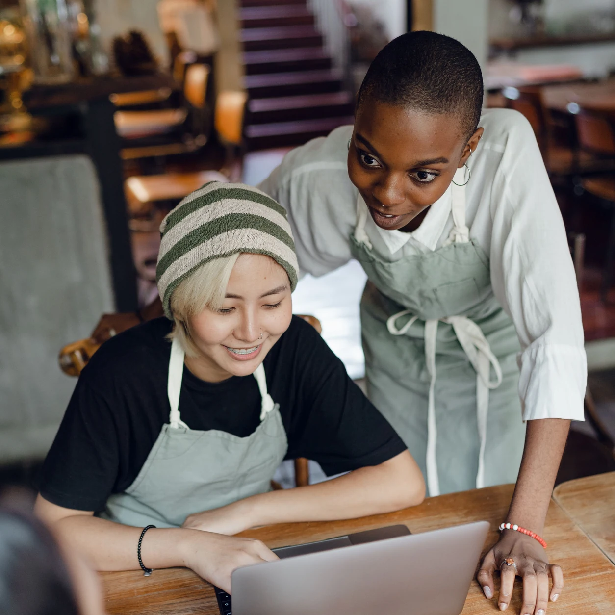 Two restaurant colleagues in aprons review a laptop together at a café table.