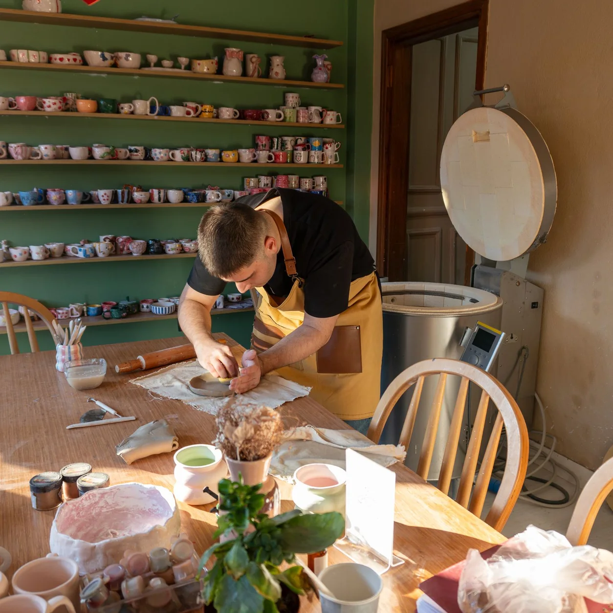 A ceramic artist at work in a sunlit studio with colorful mugs.