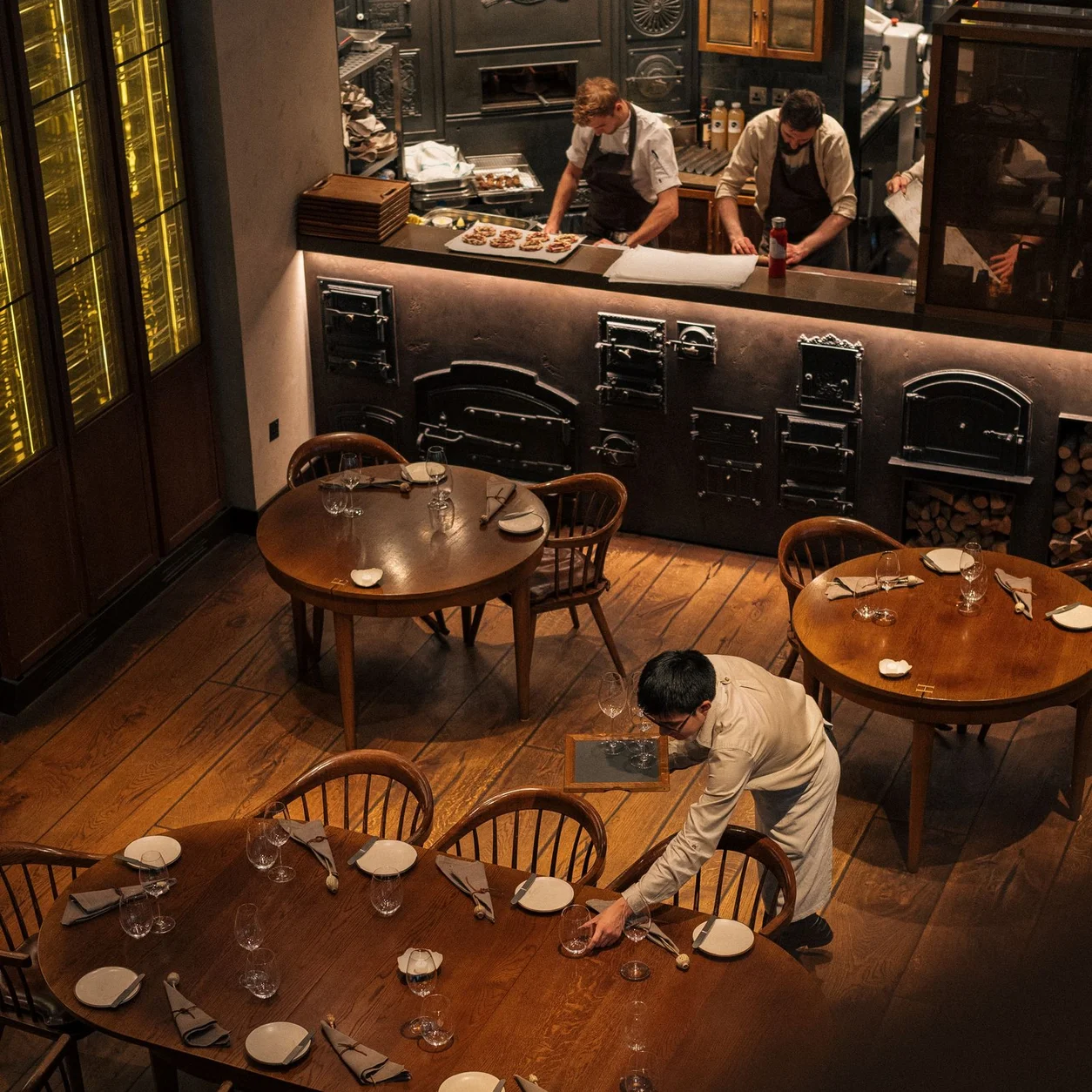 Chefs working in an open kitchen while a waiter prepares the dining room in a London restaurant.