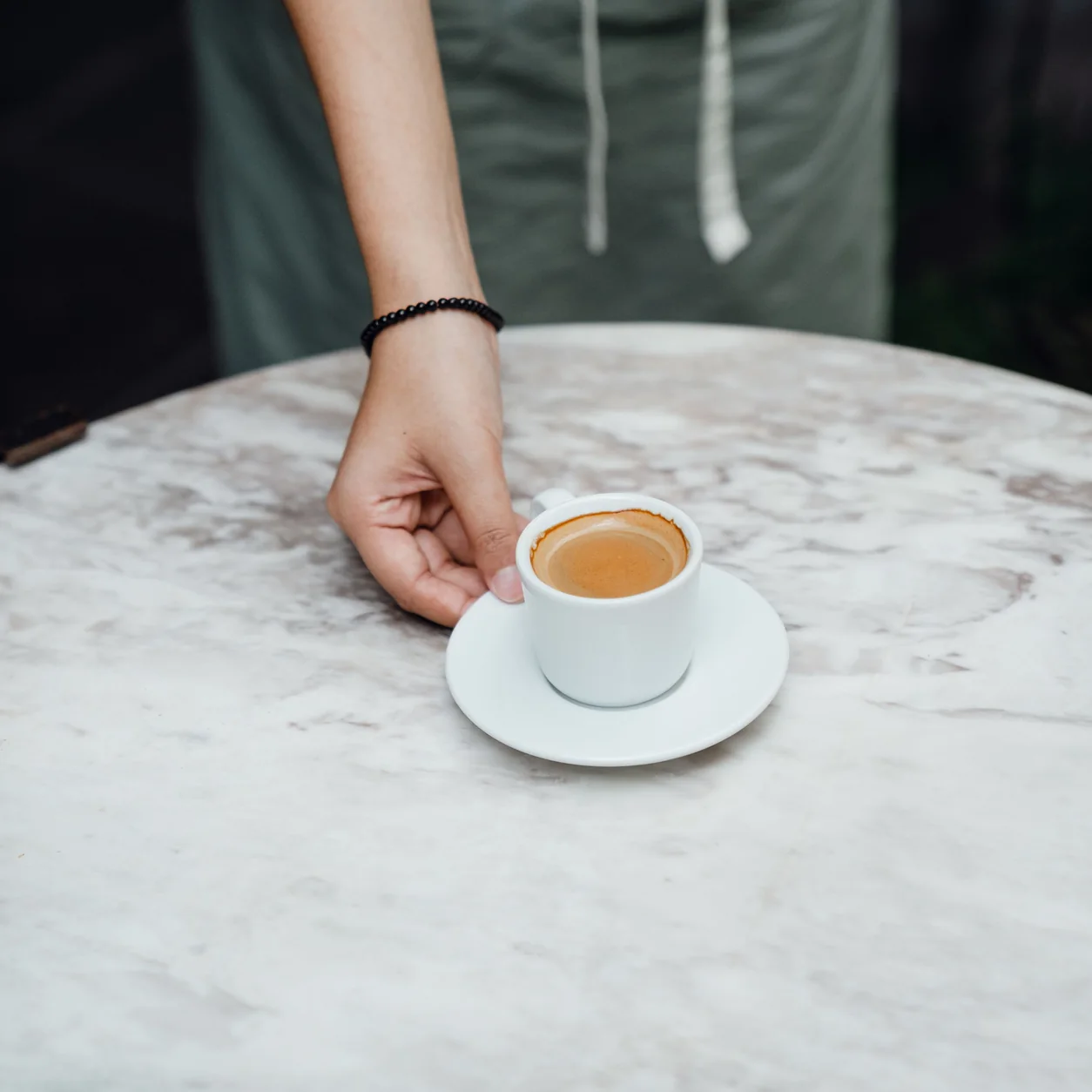 A barista in a mint apron placing espresso on a marble cafe table.