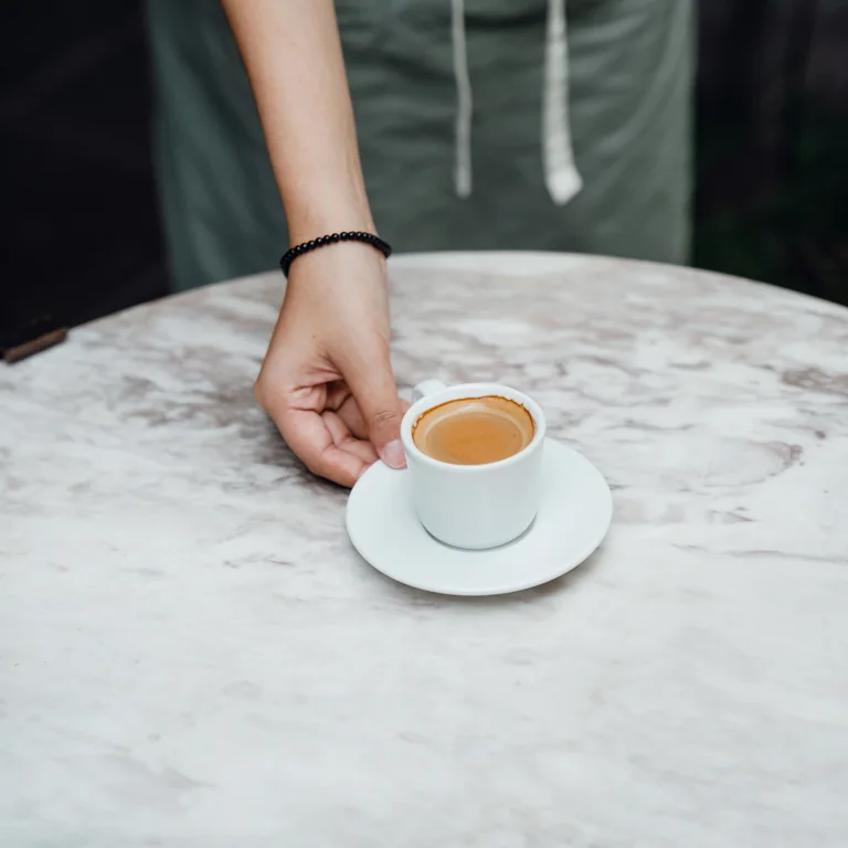 A barista in a mint apron placing espresso on a marble cafe table.
