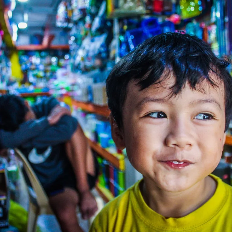 A happy child wearing a yellow shirt smiles while exploring a colorful toy store aisle, filled with various toys.
