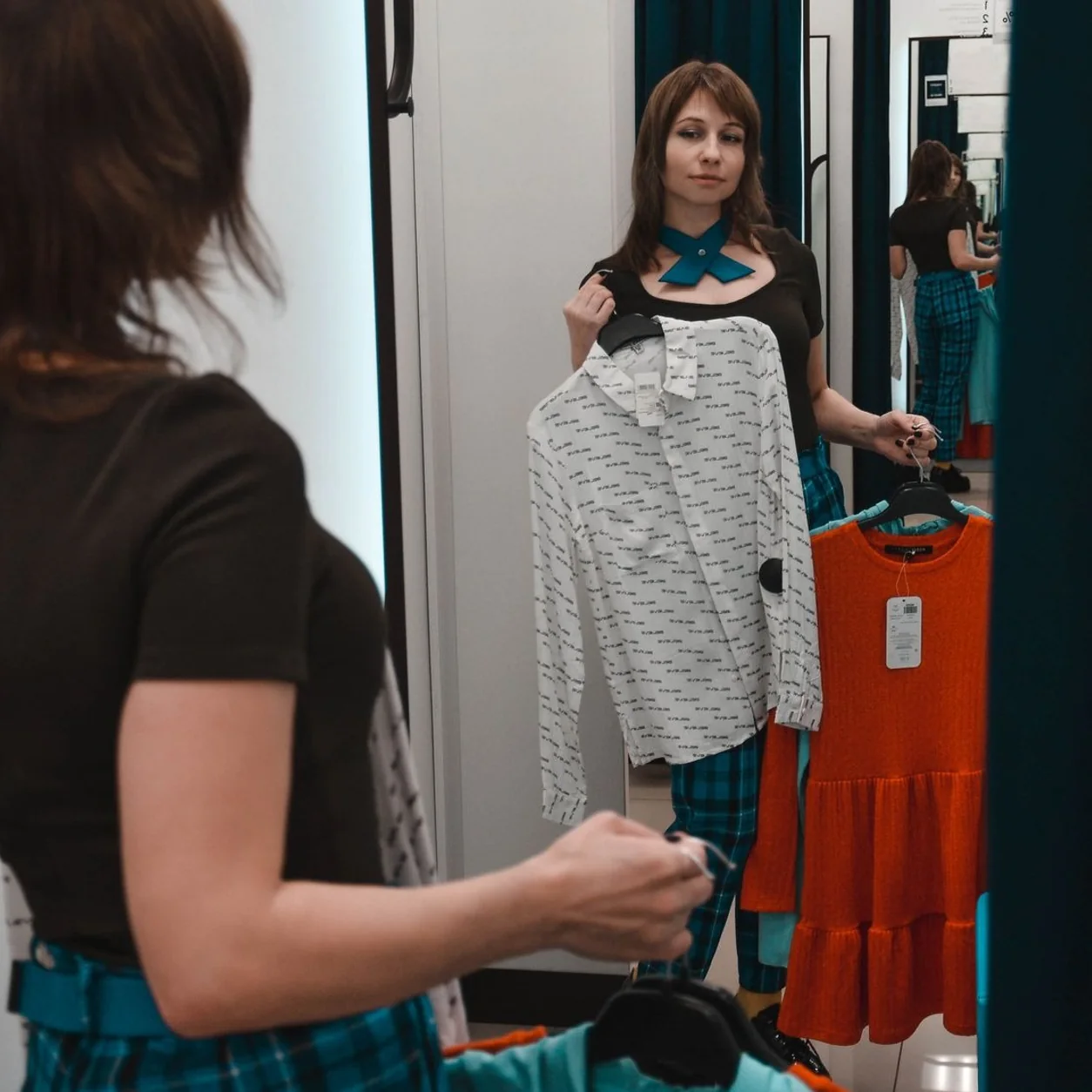 Back view of young woman standing in front of mirror in changing room of store and picking shirts
