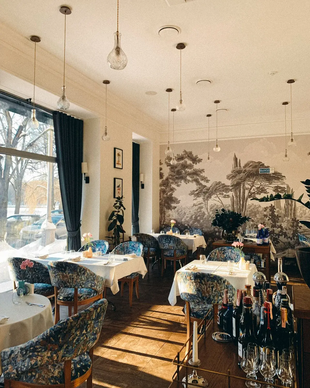 A sunlit restaurant with set tables, patterned chairs and a botanical mural.