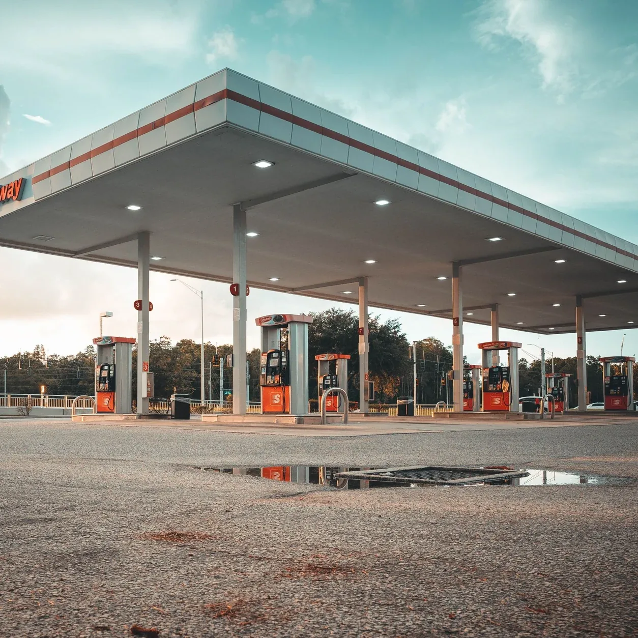 A modern fuel station with an angular illuminated canopy and red pumps at dusk.