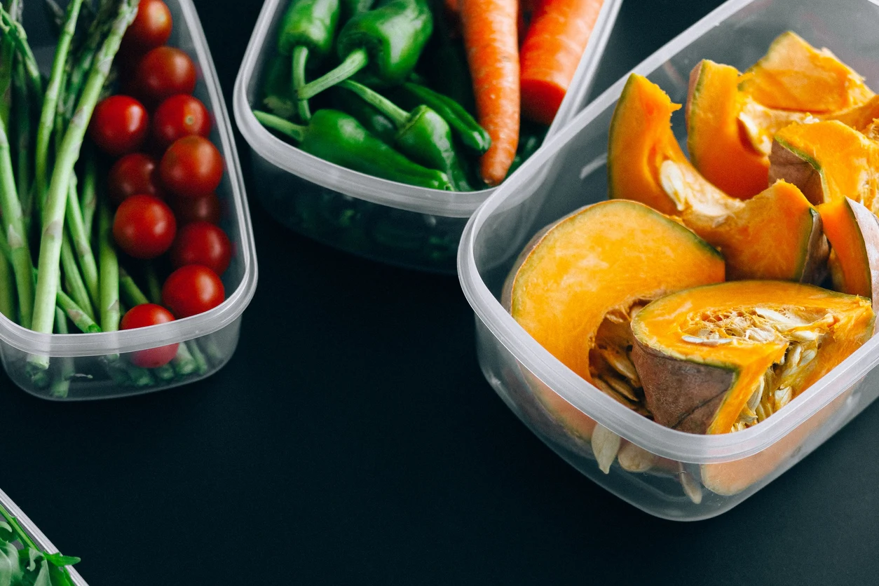 Fresh tomatoes, beans, greens and cut squash arranged in clear food containers on a dark surface.