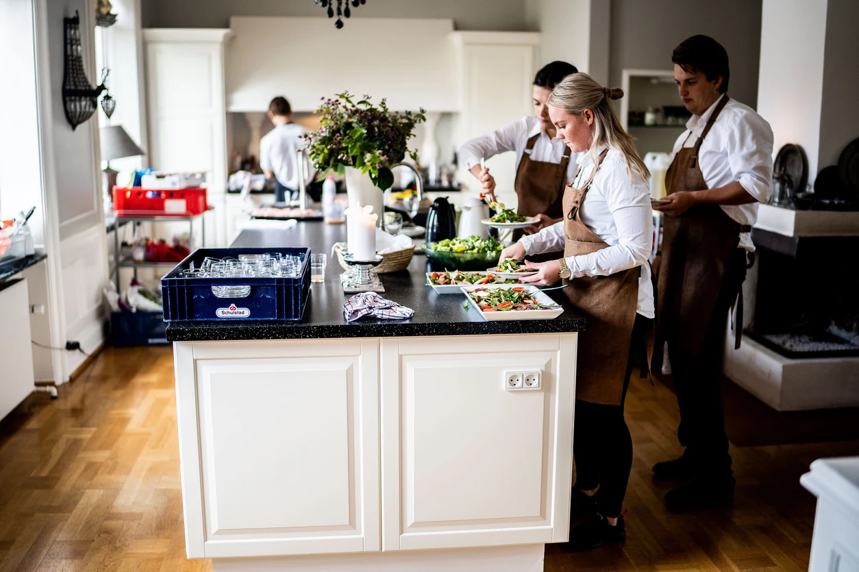 A team in white shirts and aprons prepares and arranges dishes around a kitchen island.