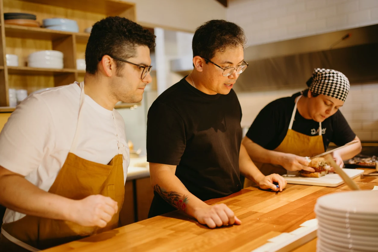 Three cooks work together at a wooden preparation counter in a restaurant kitchen.