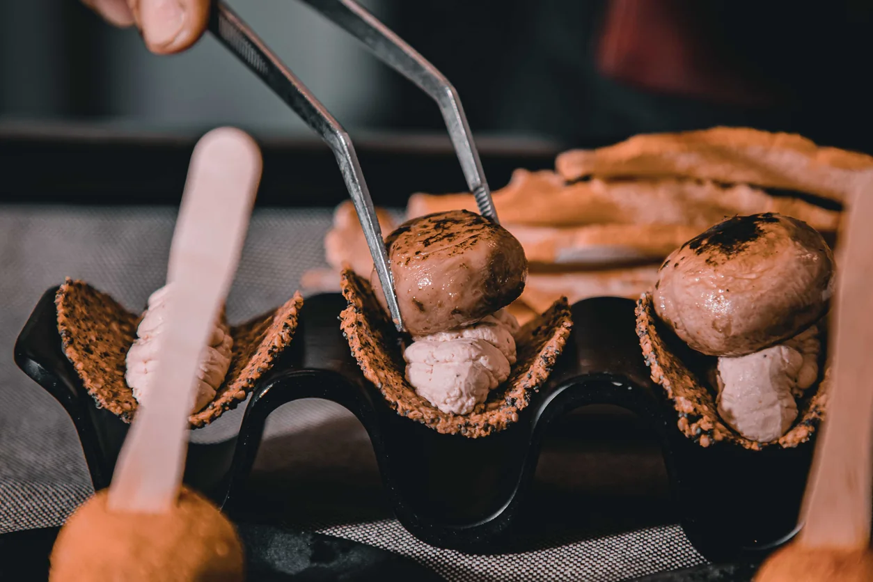 A chef uses precision tweezers to finish small mushroom-shaped appetizers on a black serving stand.