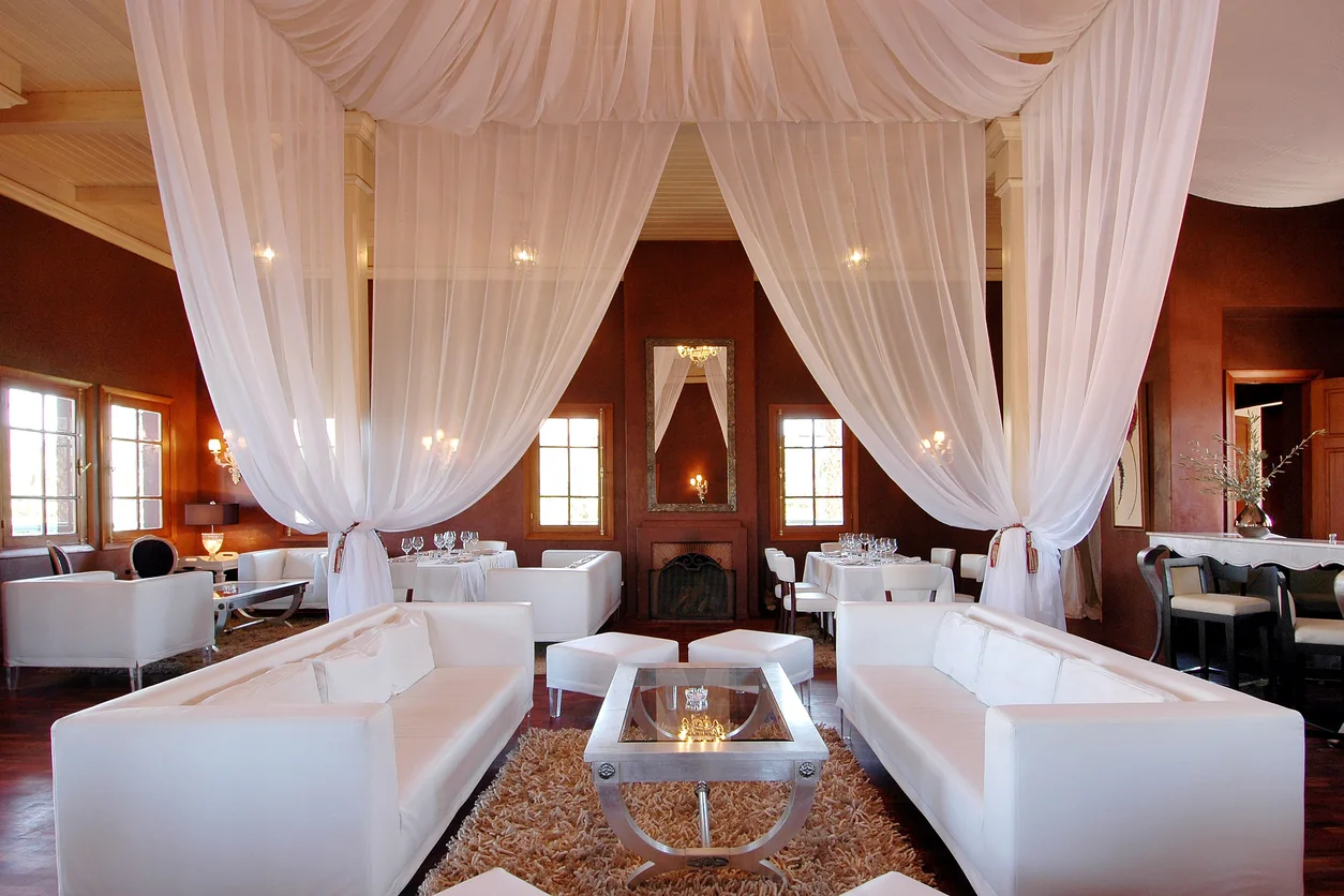 A restaurant lounge with white draped curtains, white seating and set dining tables against warm terracotta walls.