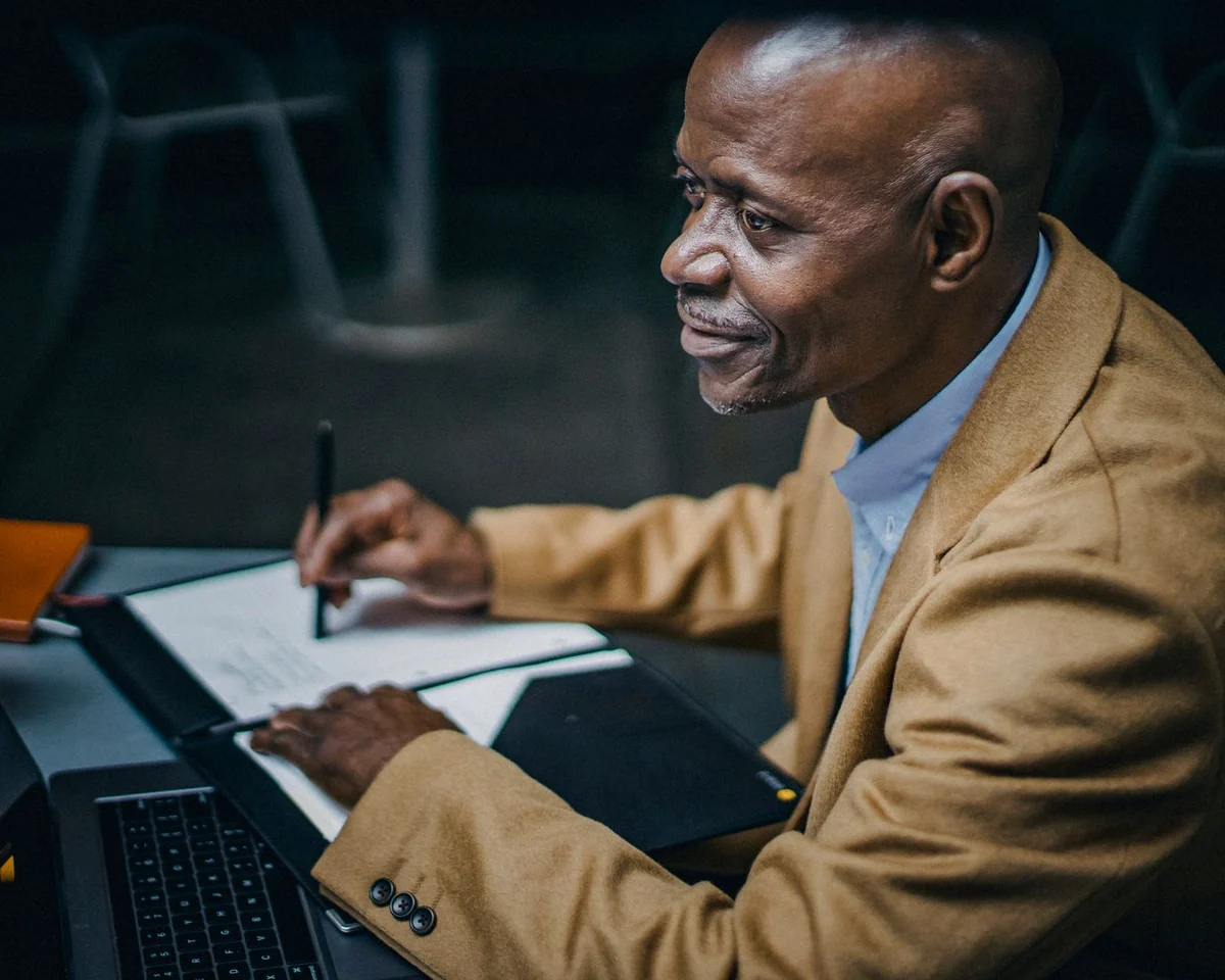 A man in a tan jacket reviewing notes beside his laptop at a cafe table.