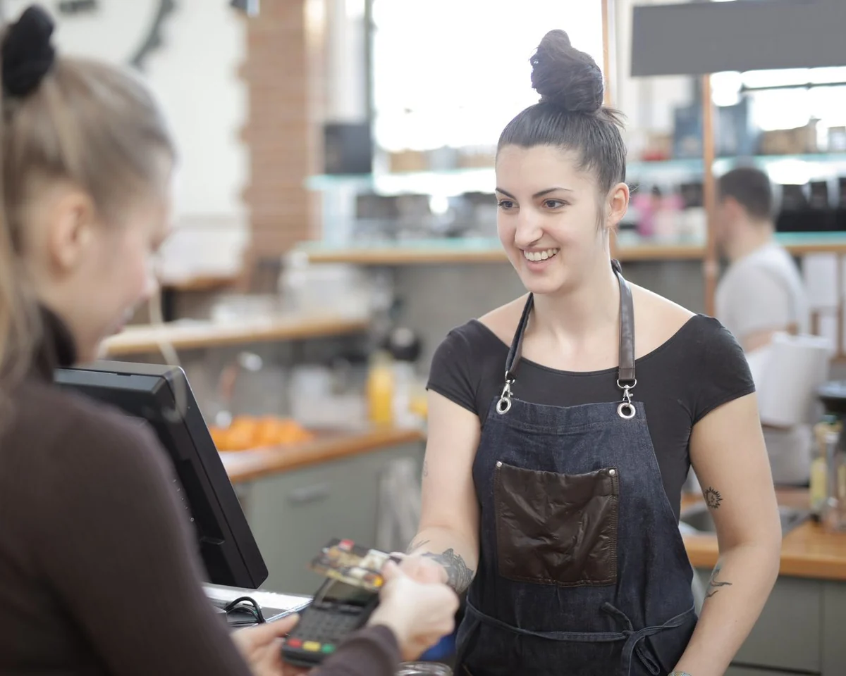 A smiling barista in a dark apron helps a guest pay at a bright cafe counter.