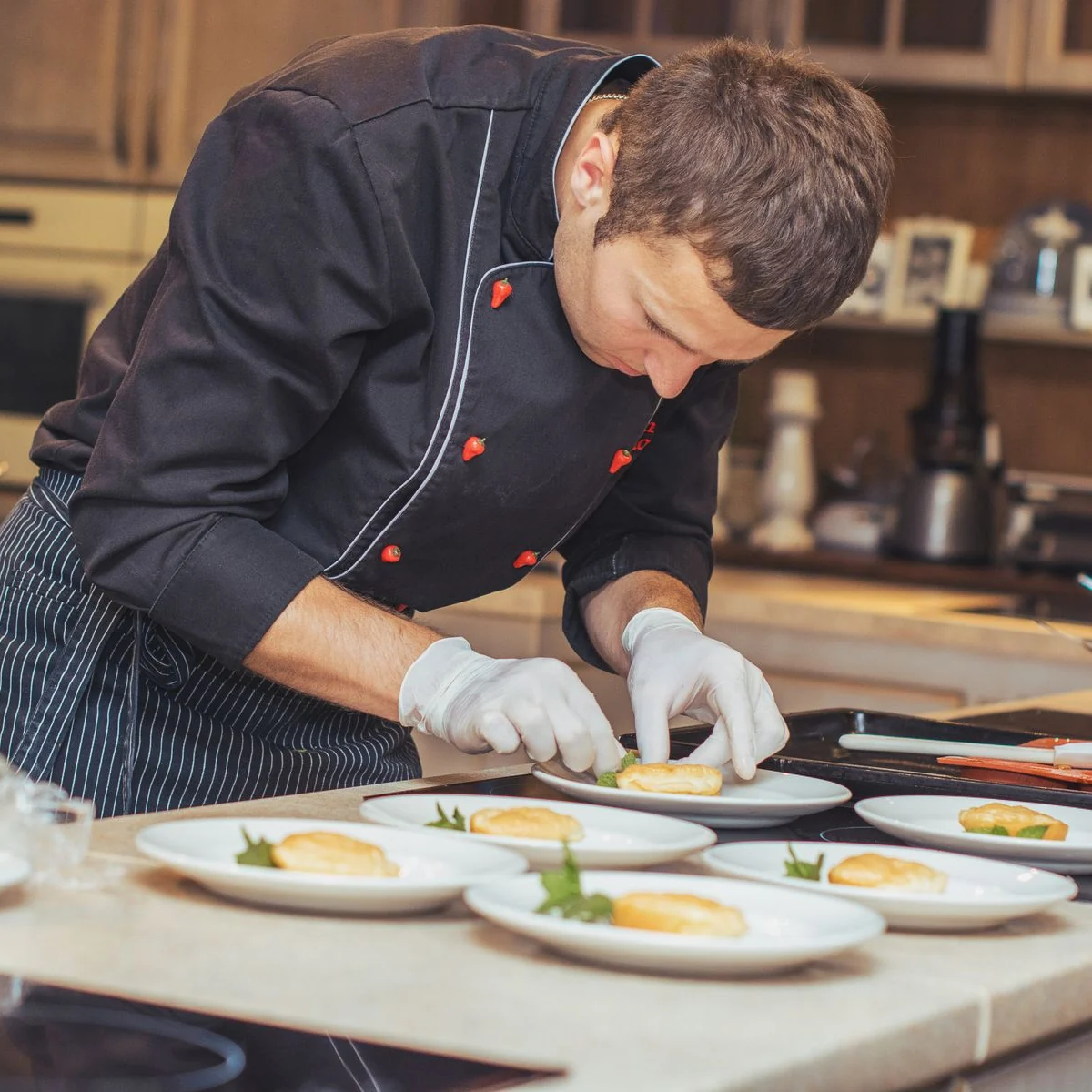 A chef in a dark jacket carefully plating dishes in a warm, wood-lined professional kitchen.