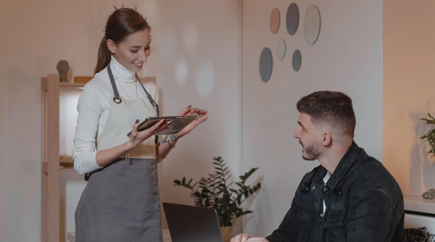 A waitress takes an order on a tablet beside a café guest.