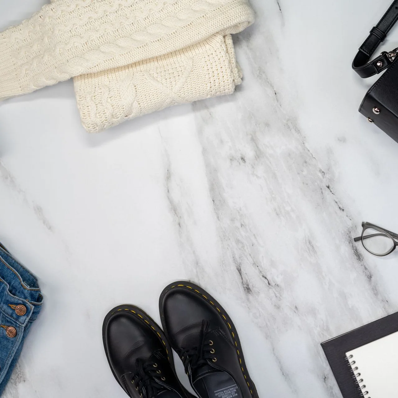 A cream knit sweater, denim, black shoes and accessories arranged on pale marble.