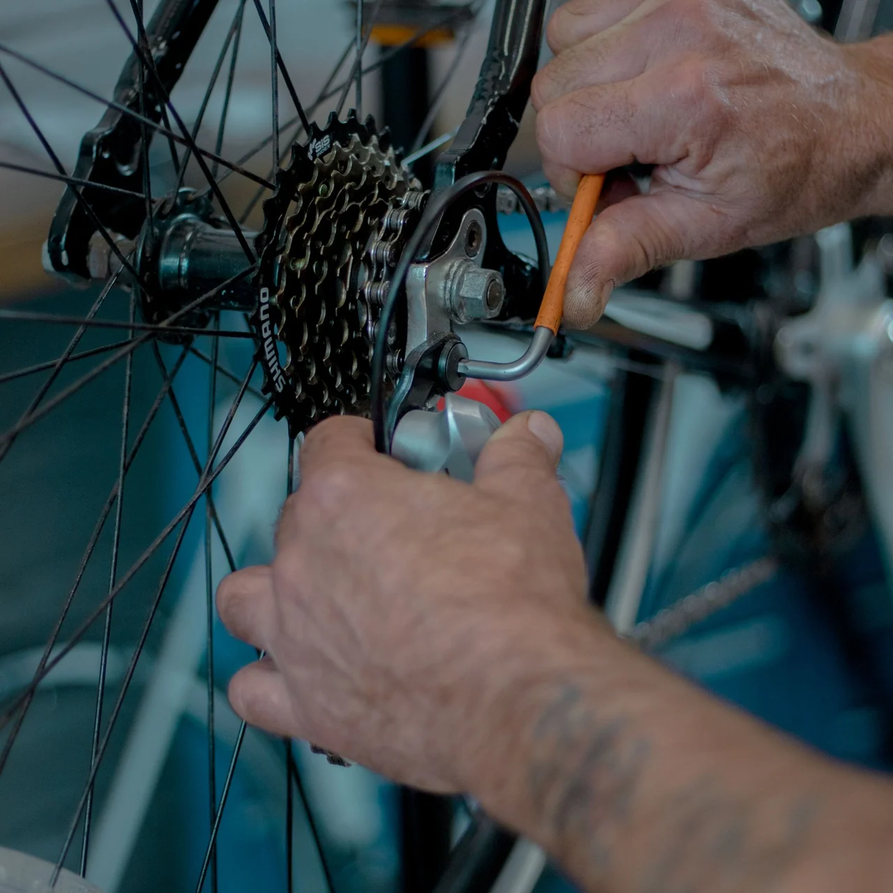 A bicycle mechanic works on a rear wheel beside a blue frame in a workshop.