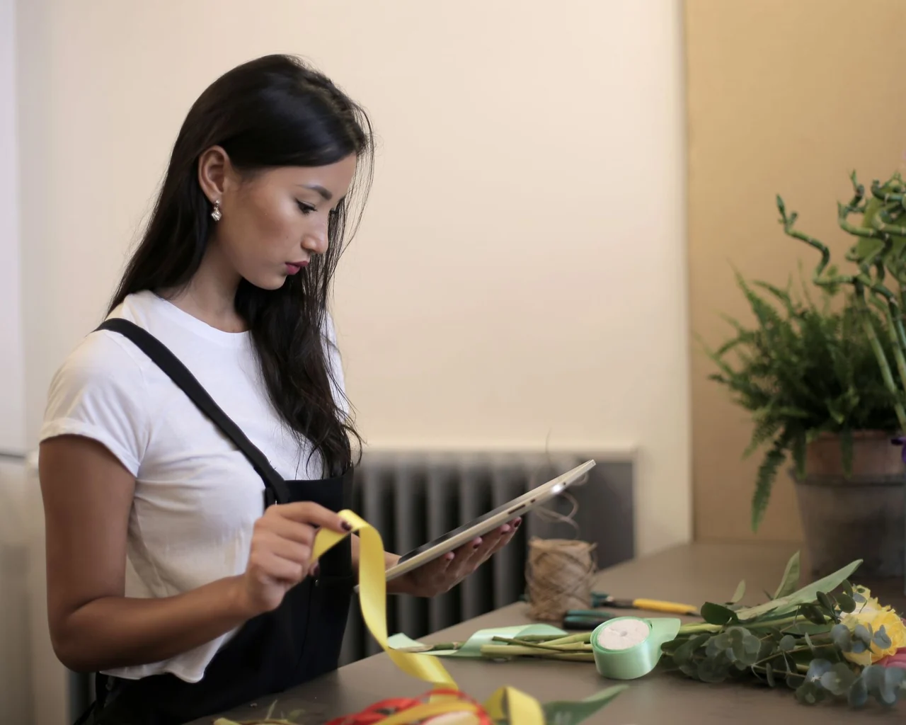 A florist in a black apron checks a tablet while preparing flowers and ribbons at her shop counter.