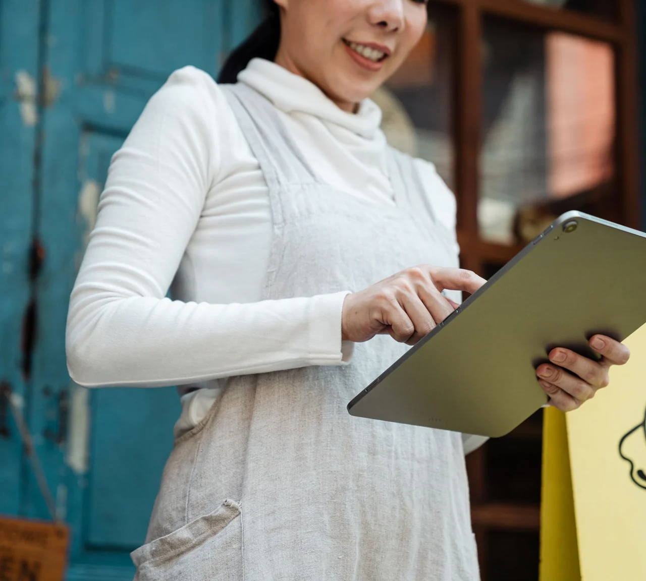 A shop owner in a white apron holds a landscape tablet at a turquoise storefront.